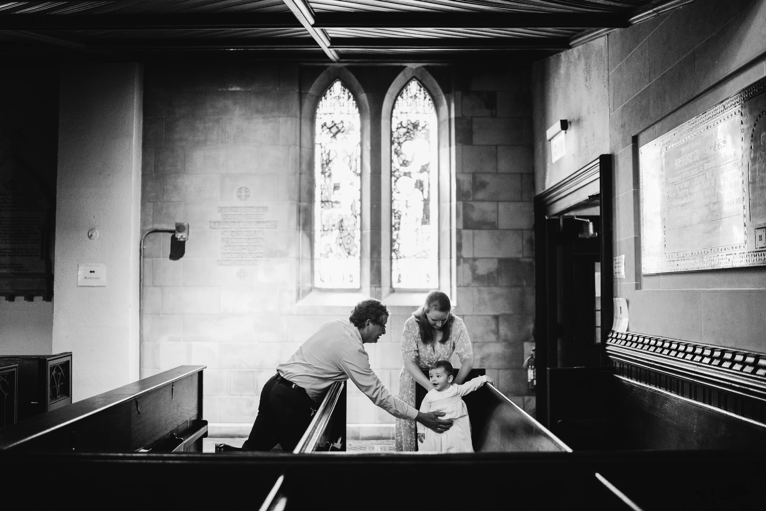Man and woman steadying a baby in a white gown between church pews beneath tall arched windows