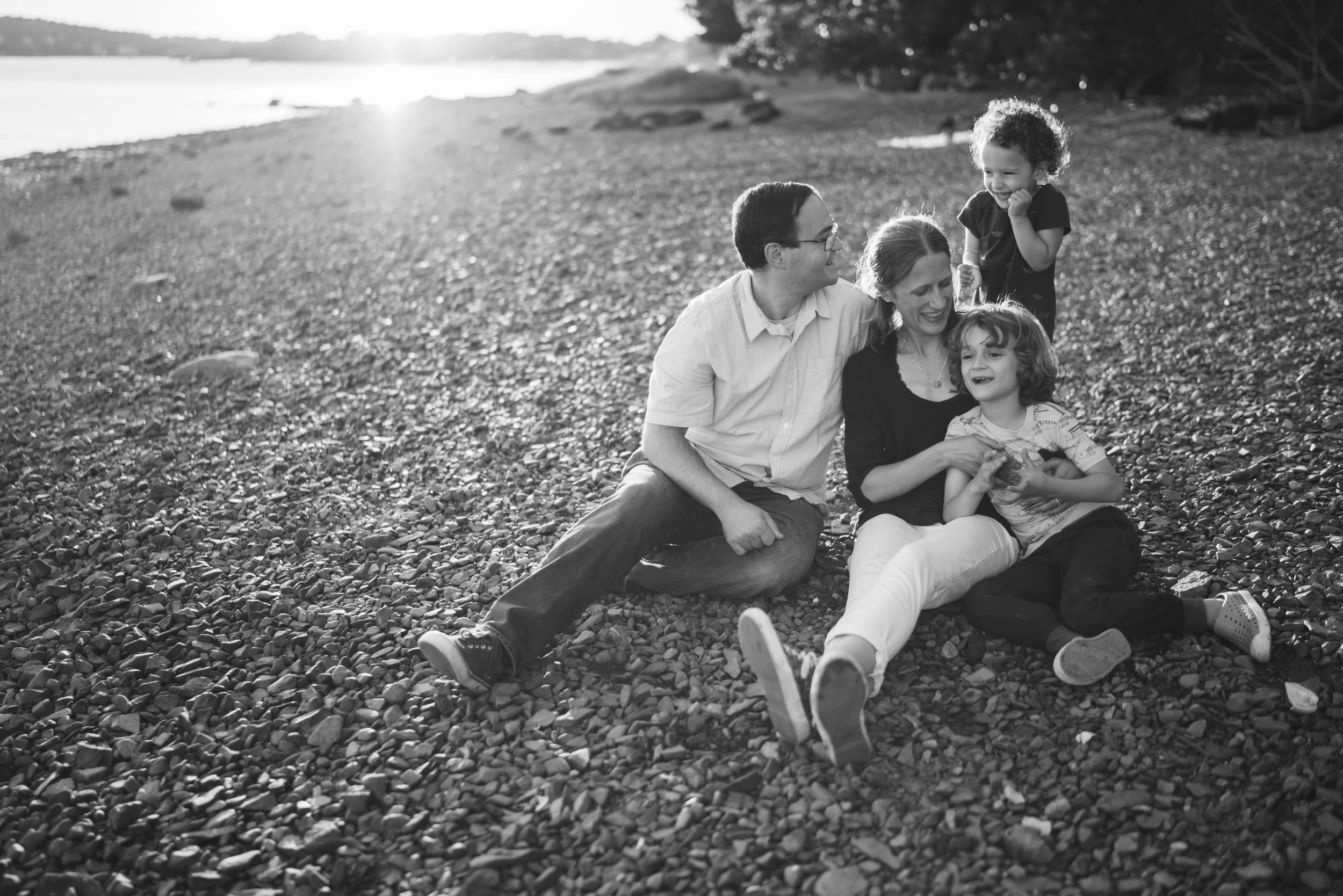 Family of four sitting together on a pebble beach, backlit by low sun over the water