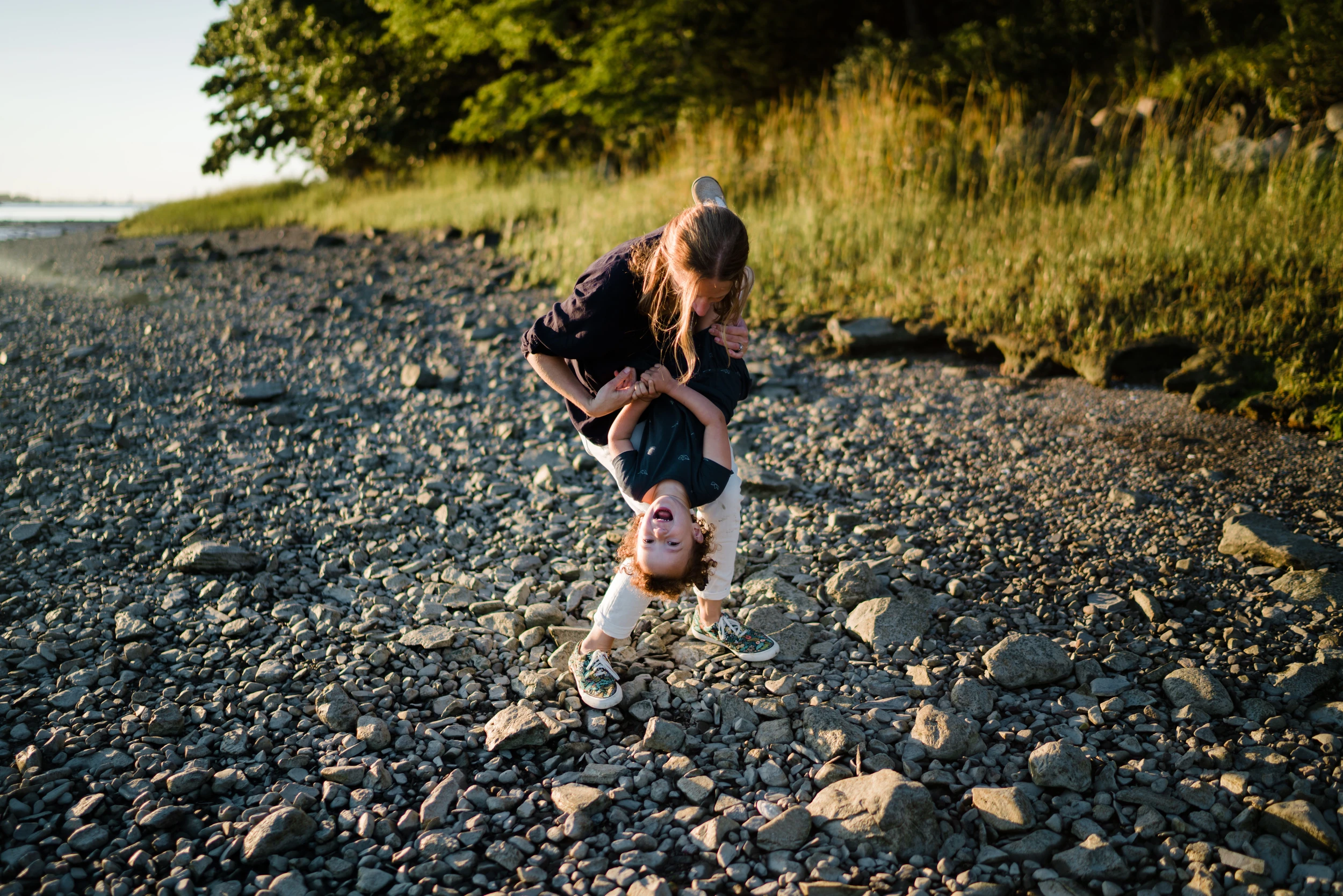 Older child holding a laughing younger child upside down on a pebble beach in low sun, grasses behind