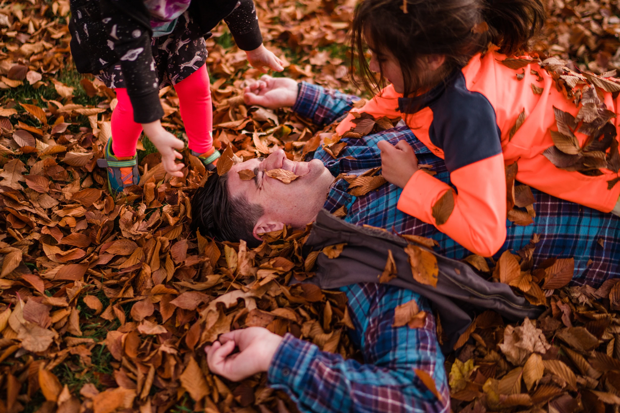 Father lying in a pile of autumn leaves while two children cover him