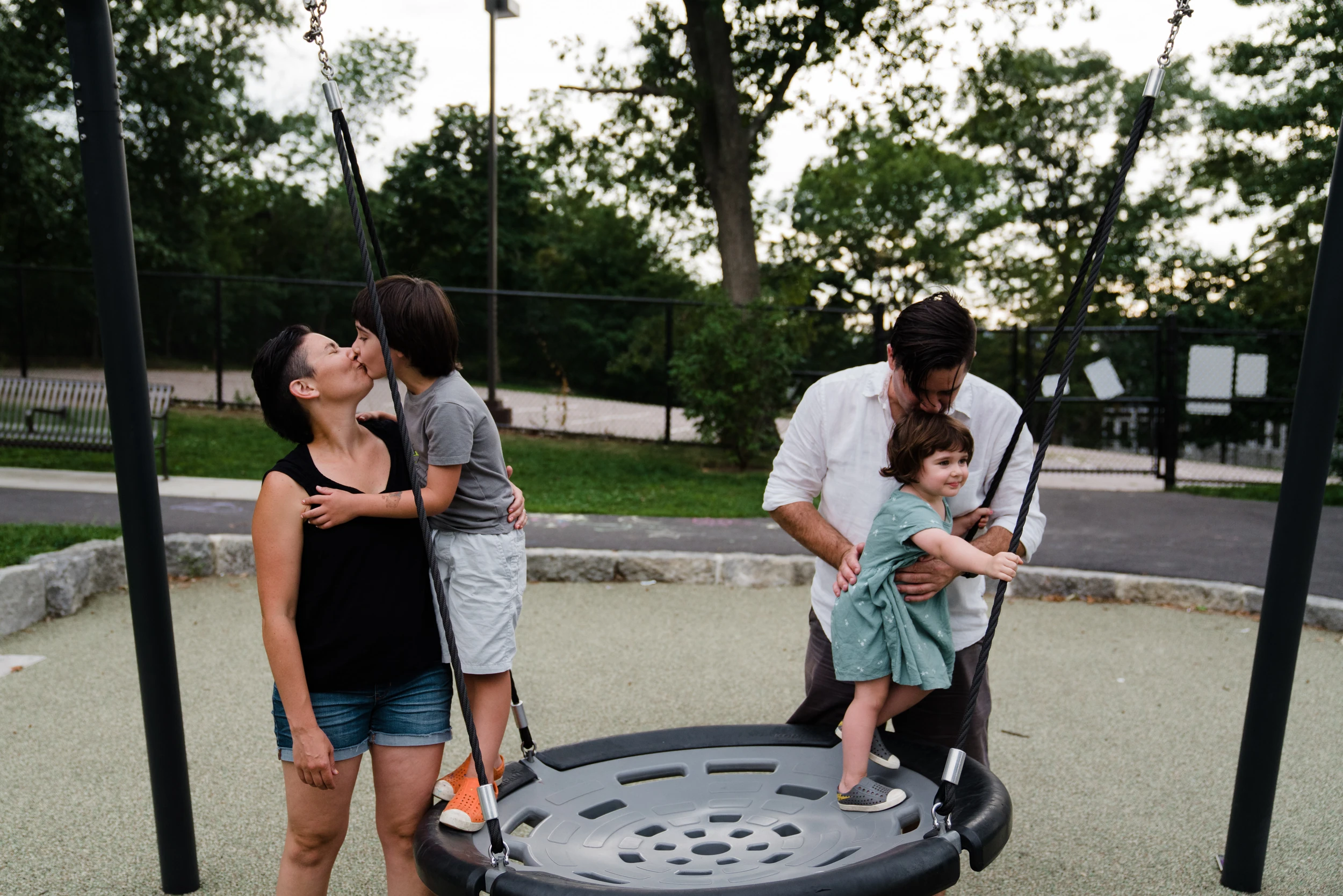 Mother kisses a son standing on a nest swing while the father holds a daughter on it, park at dusk