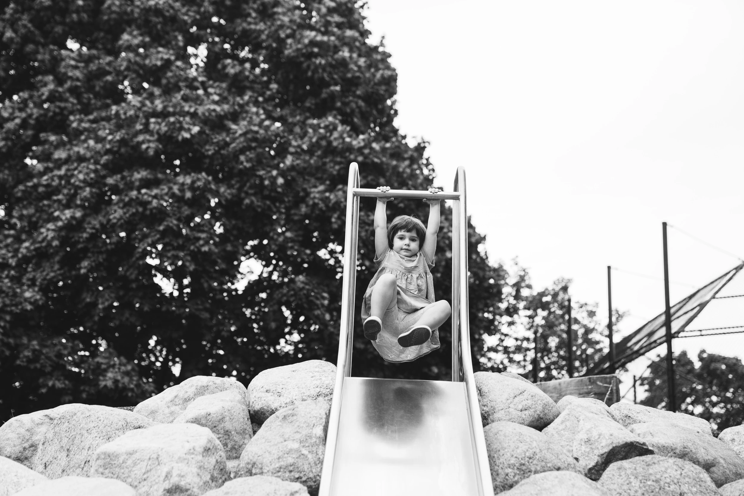 Girl hanging from the bar at the top of a metal slide set into a pile of boulders, large tree and white sky behind