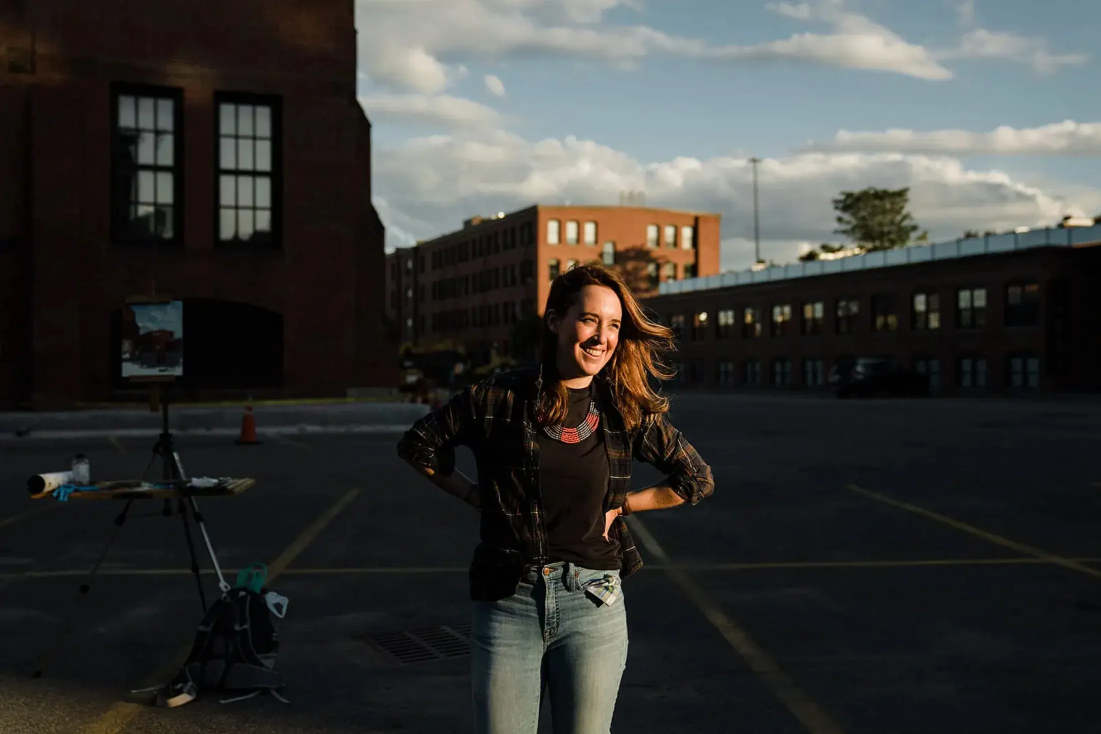 Woman smiling with hands on hips in an empty lot at golden hour, brick mill buildings and sky behind