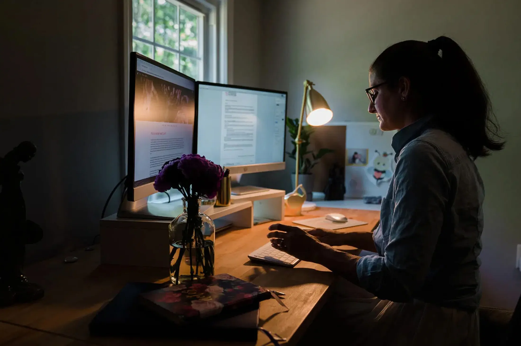 Woman typing at a desk with two monitors in a dim room, a desk lamp lit