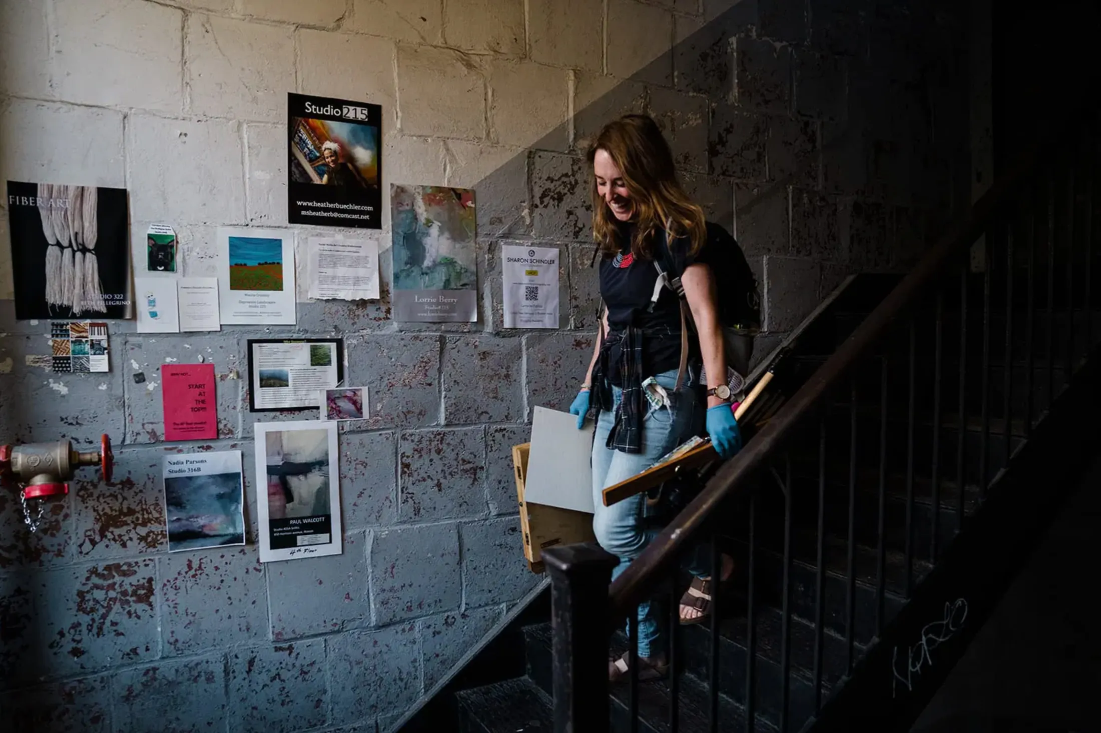 Woman with papers and gloves walking down a stairwell beside a cinderblock wall of posters