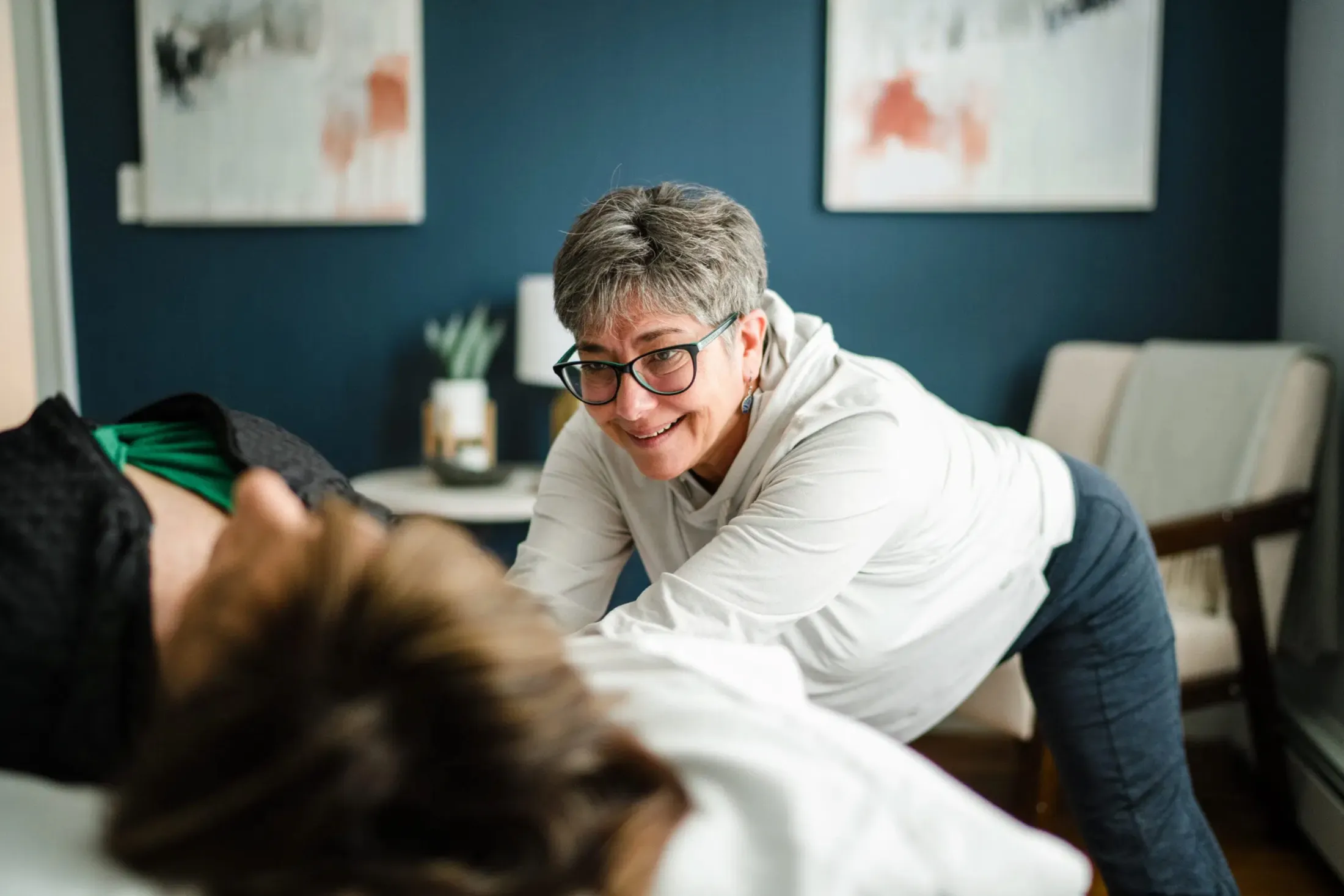 Massage therapist smiling as she leans over a client in a room with blue walls and paintings