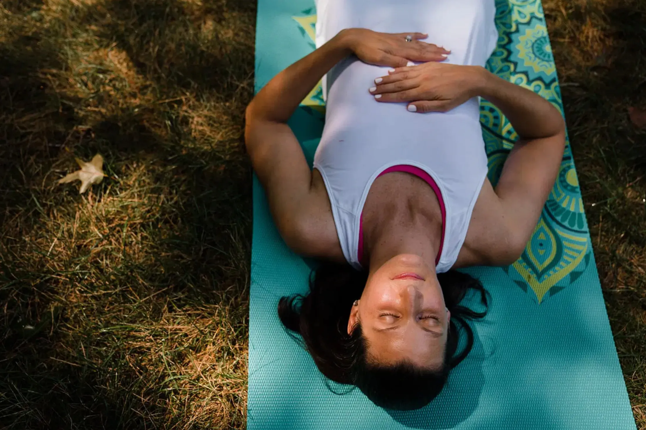 Woman lying on a teal yoga mat on the grass, seen from above, hands on her chest