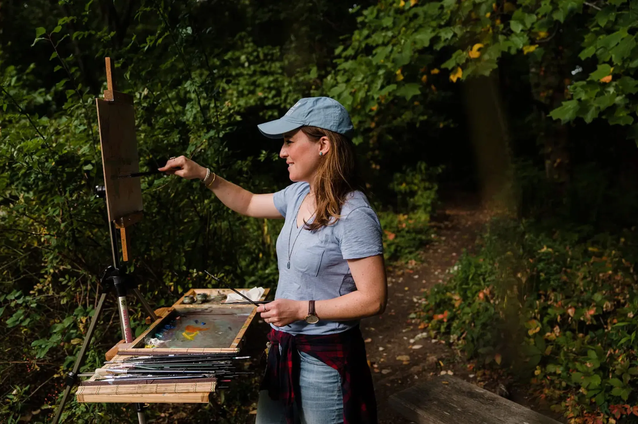 Woman in a blue cap painting at an outdoor easel in the woods