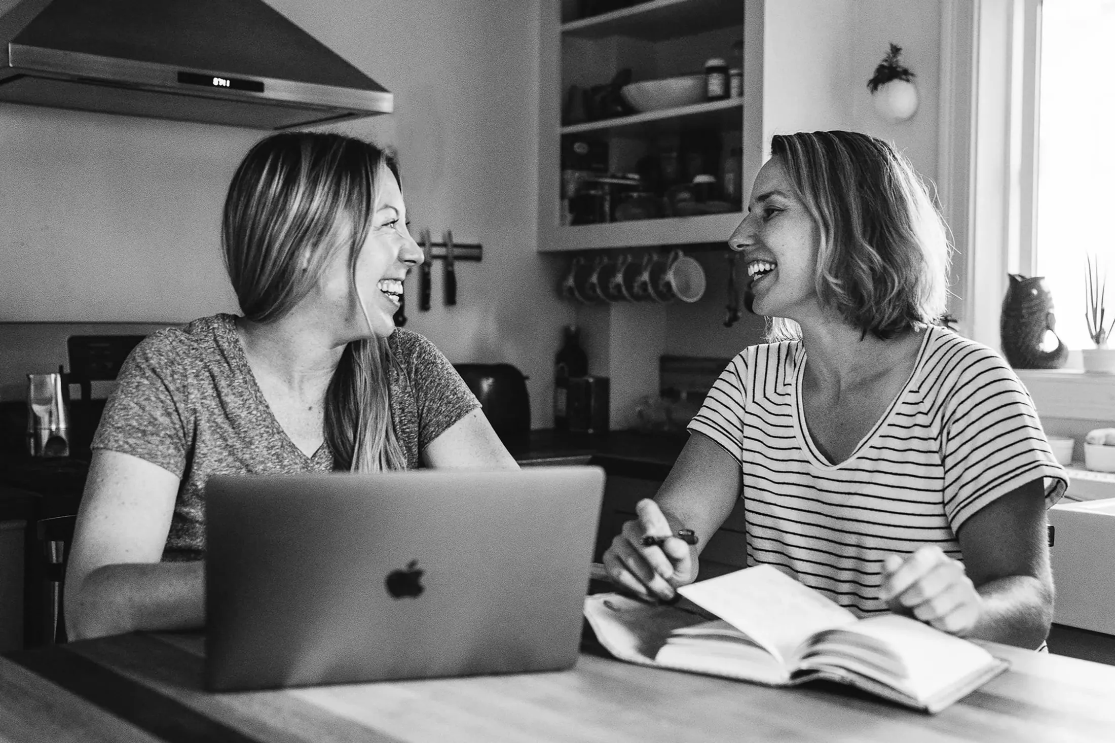 Two women laughing at a kitchen table over a laptop and a notebook