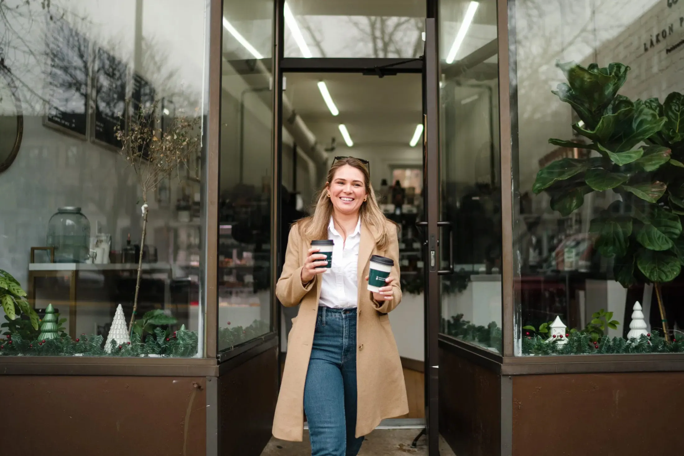 Woman in a camel coat walking out of a glass shop door holding two coffees