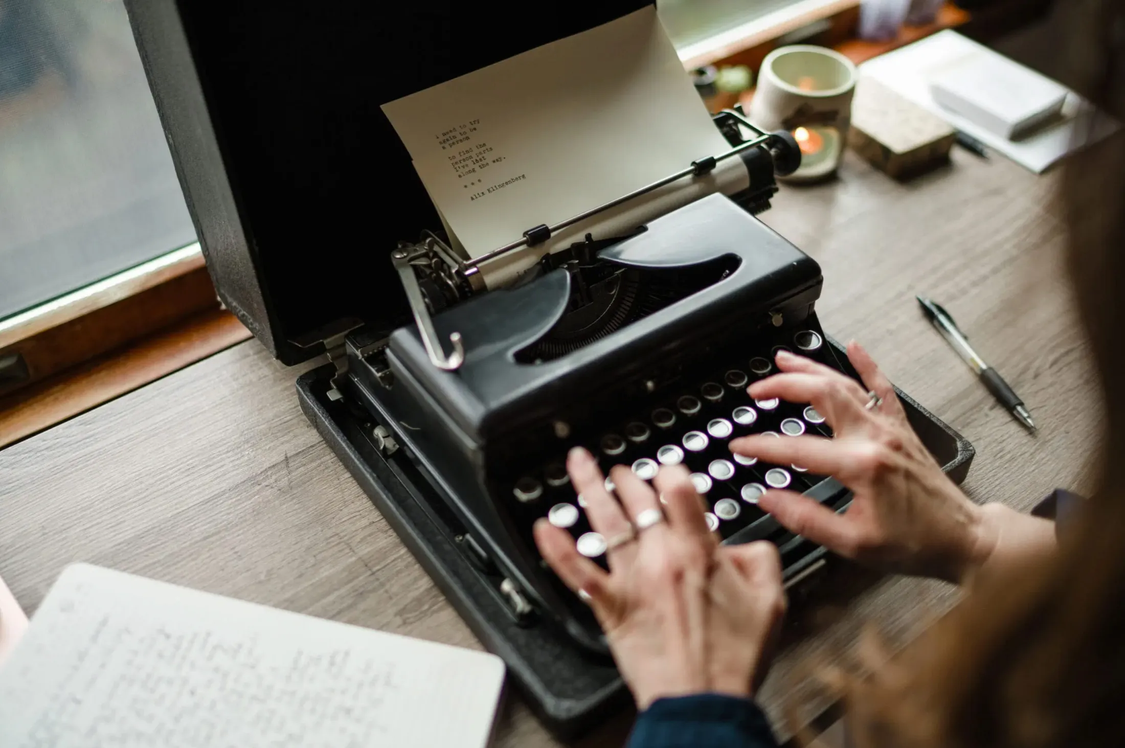 Hands typing on a vintage typewriter beside notebooks and a mug