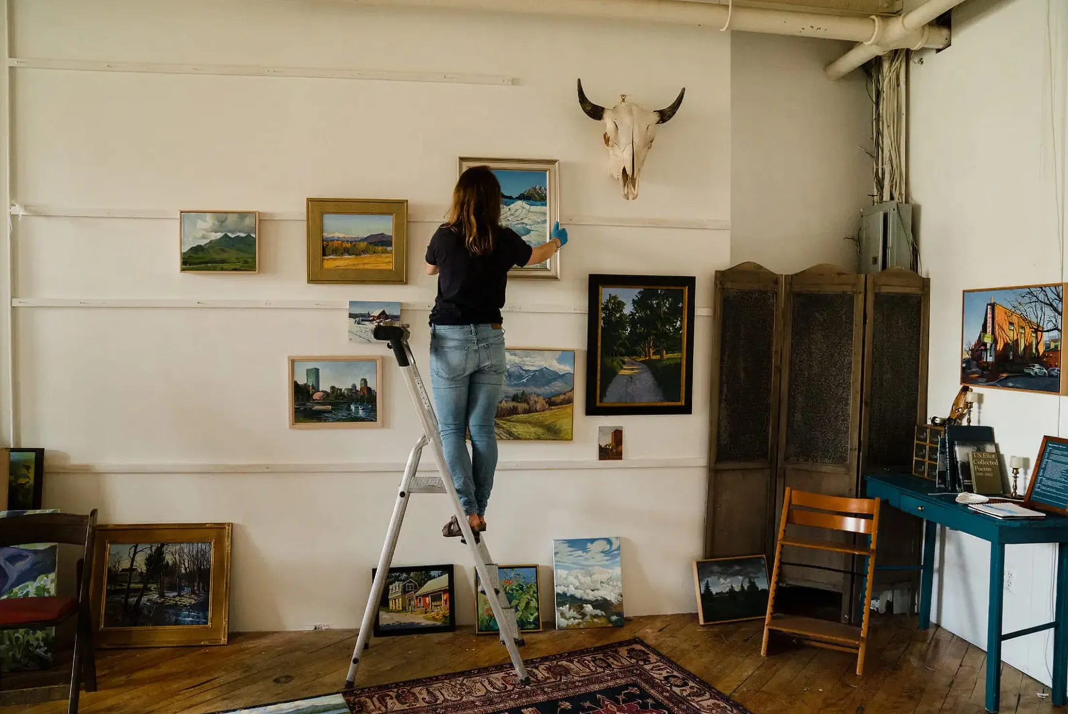 Woman on a stepladder hanging landscape paintings on a white gallery wall