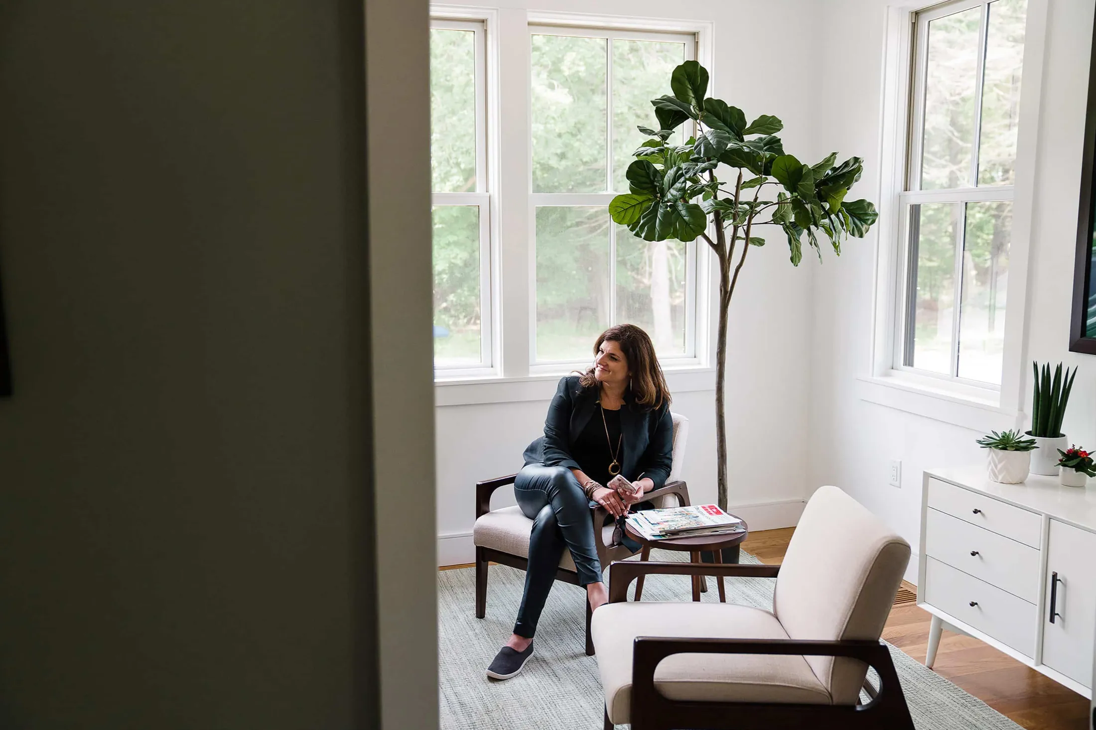 Woman sitting in an armchair by tall windows and a fig tree, seen through a doorway