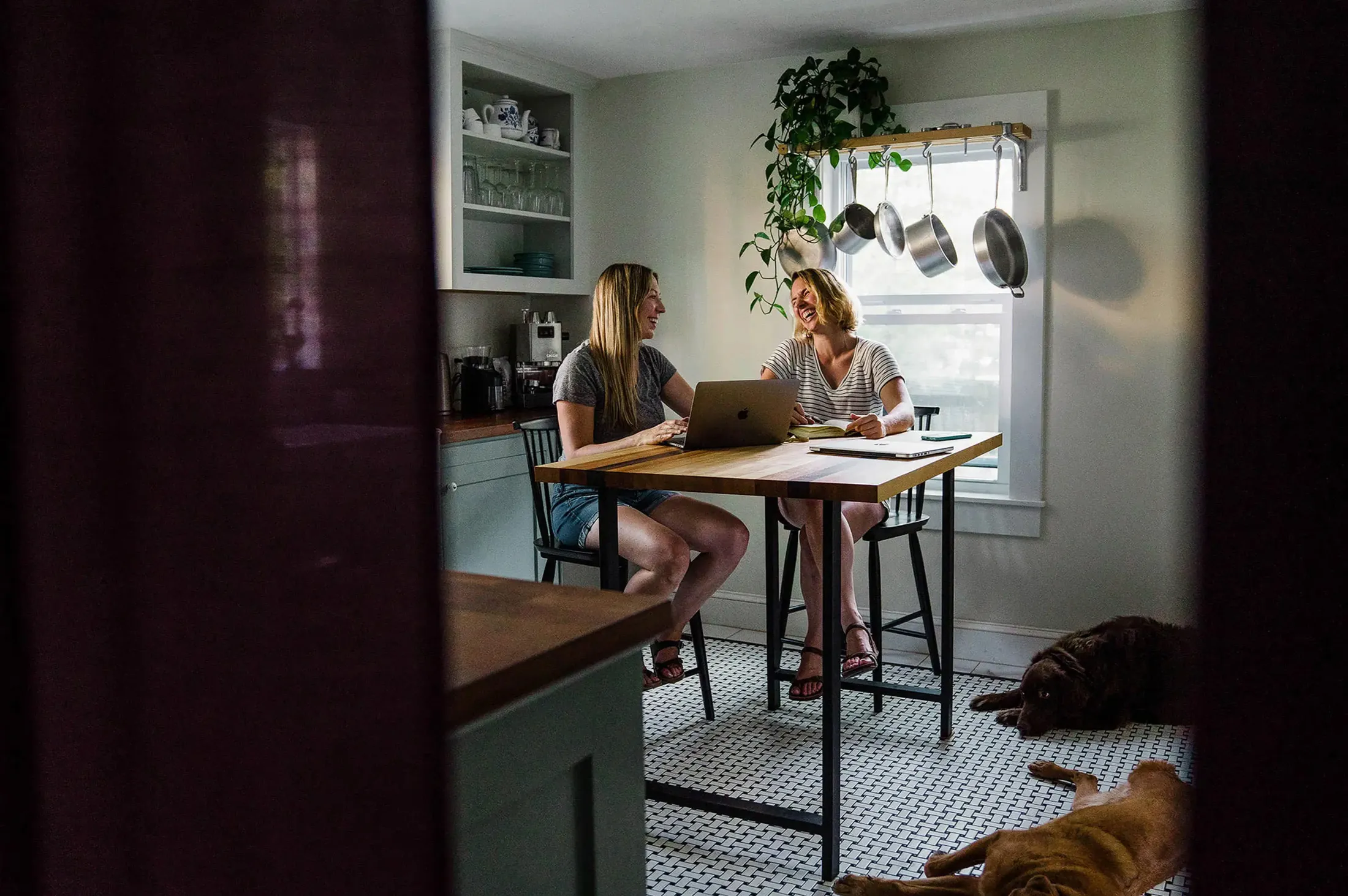 Two women working at a kitchen table with a laptop, a dog on the floor, framed by a dark doorway