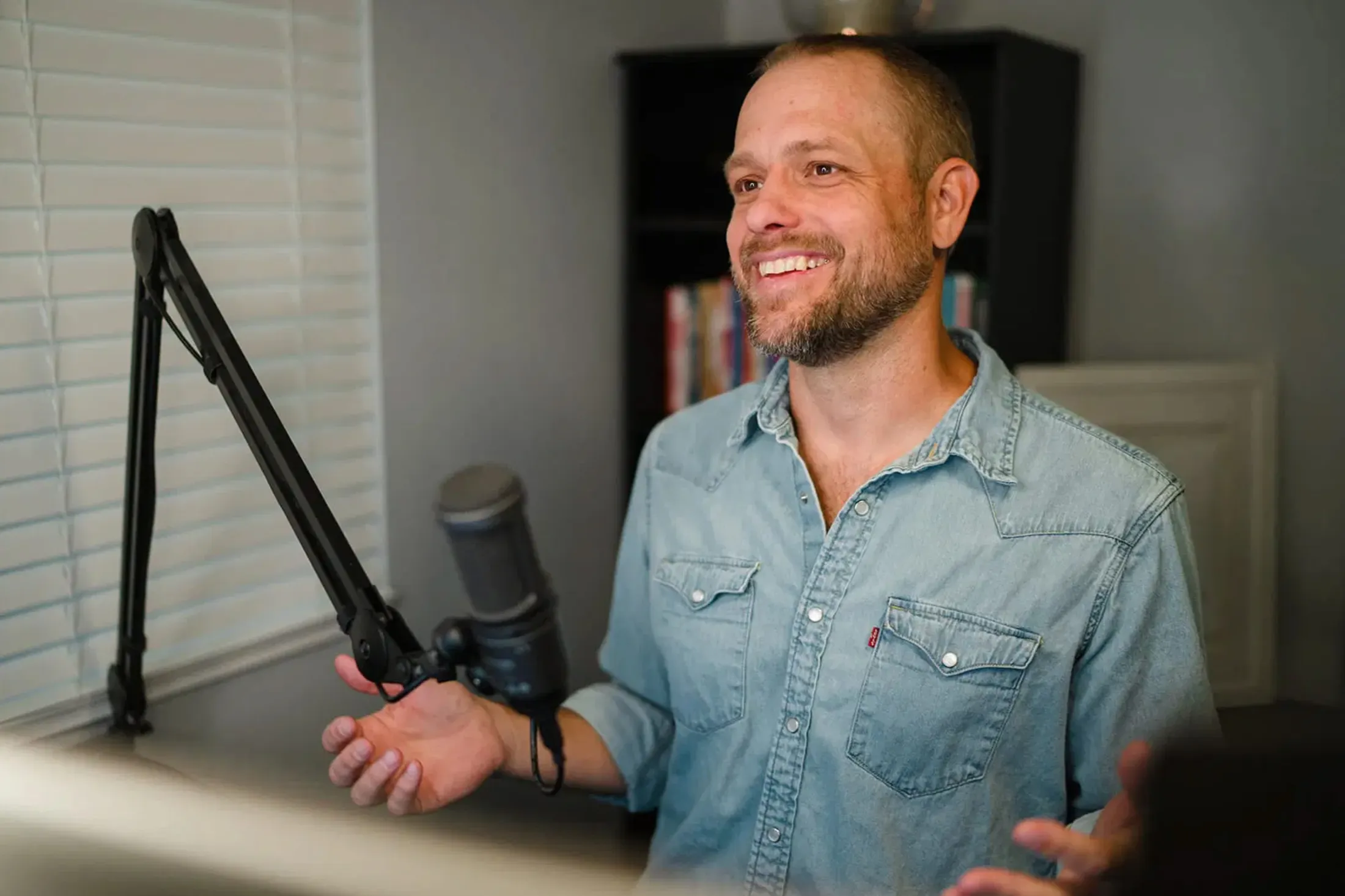 Bearded man in a denim shirt laughing beside a podcast microphone