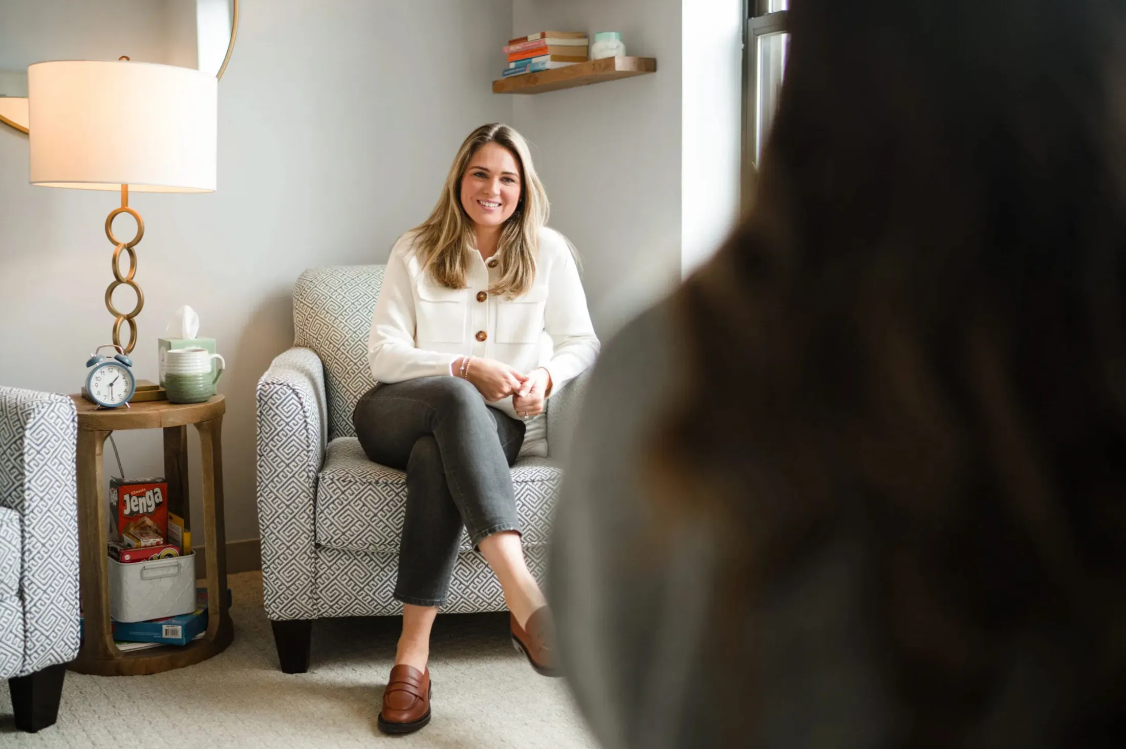 Woman in a cream sweater laughing in an armchair, seen past someone's shoulder
