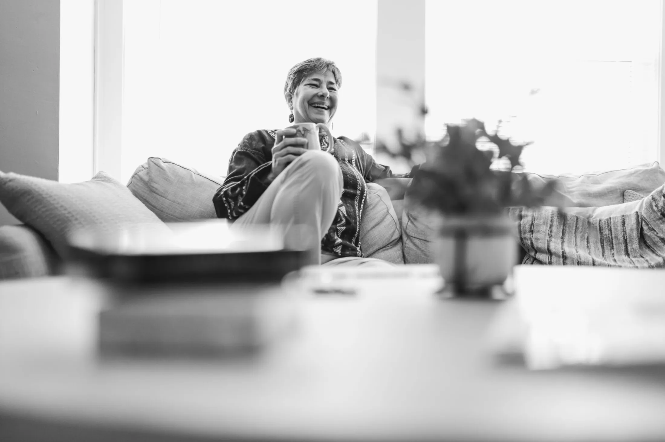 Older woman laughing on a sofa holding a mug, a table soft in the foreground