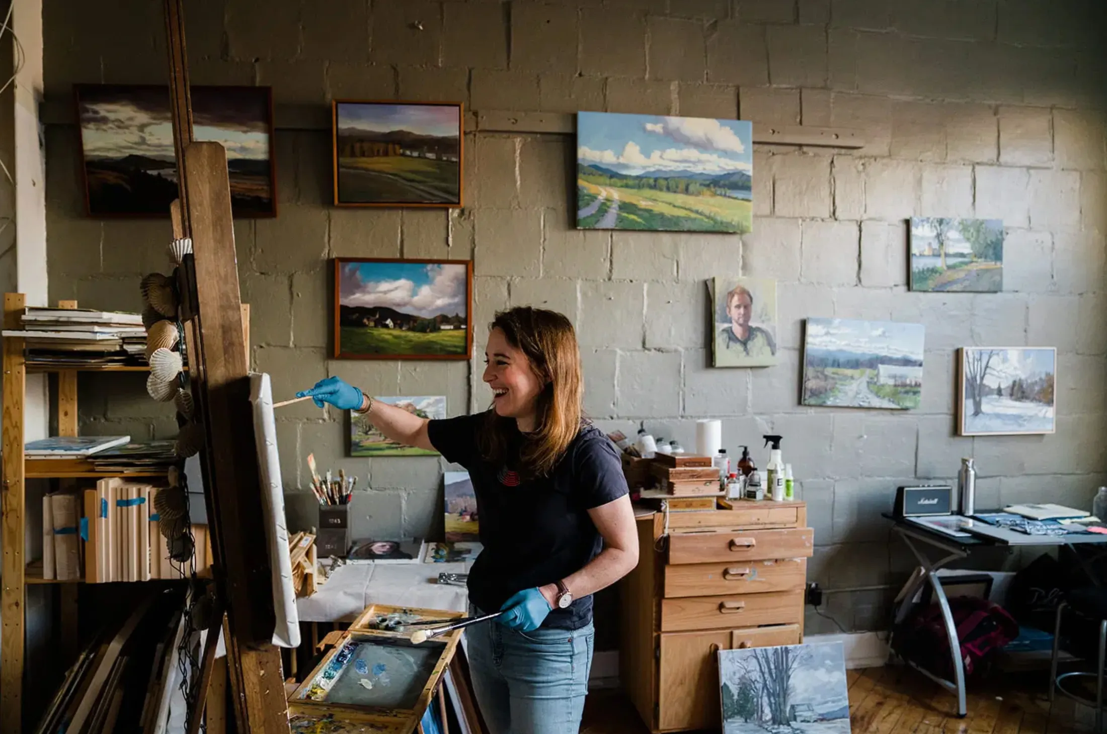 Painter smiling at her easel in a studio hung with landscape paintings