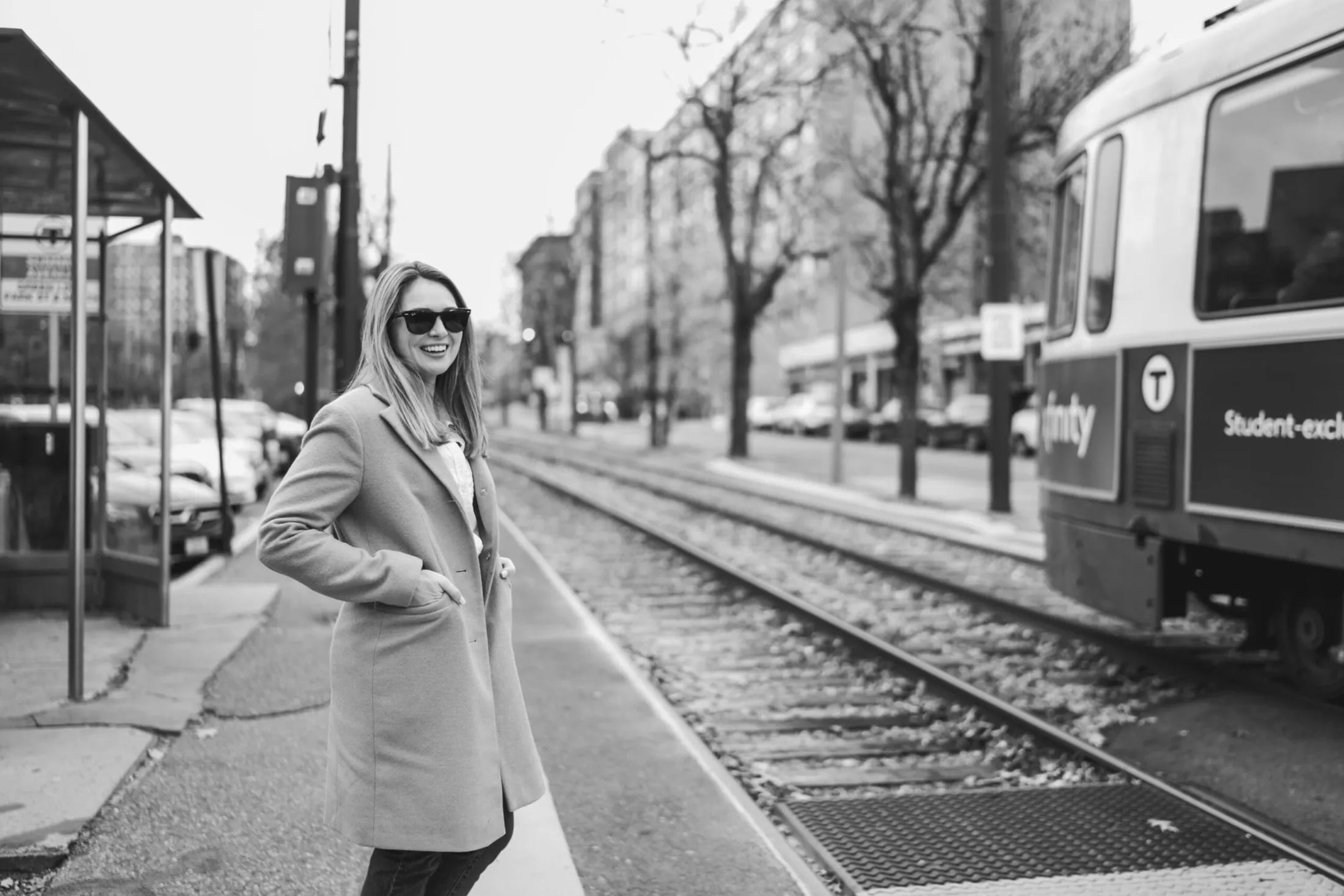 Woman in a long coat and sunglasses on a sidewalk beside tram tracks and a trolley
