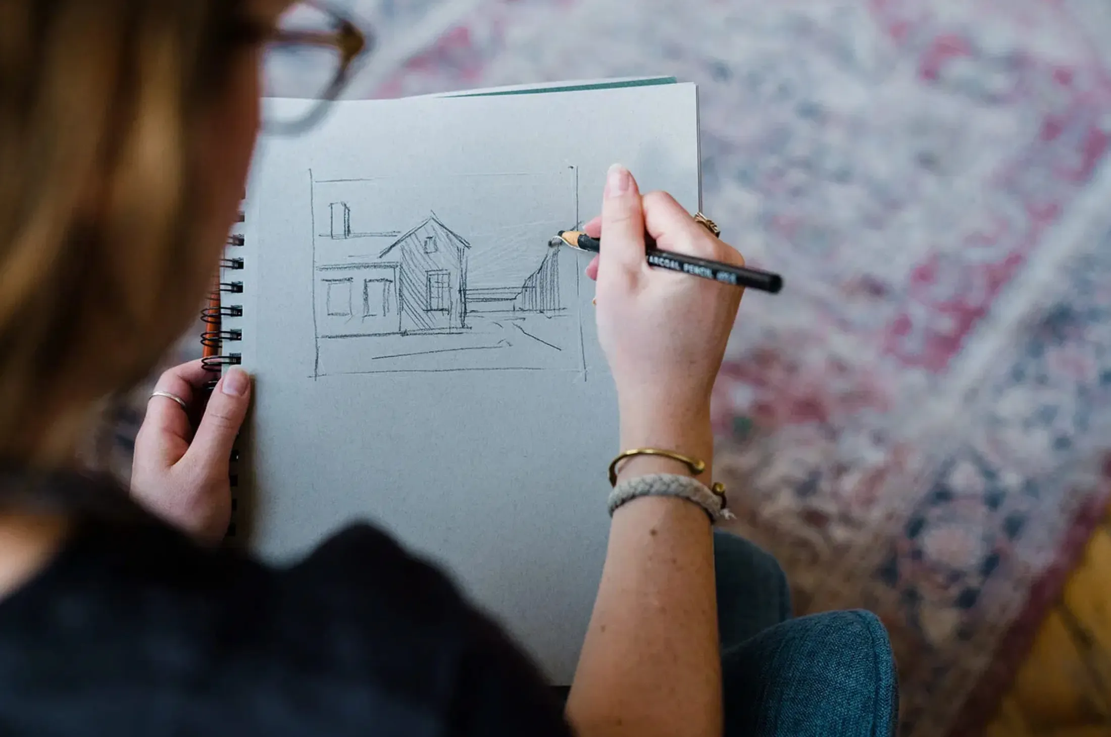 Hand sketching a house in a sketchbook over a patterned rug