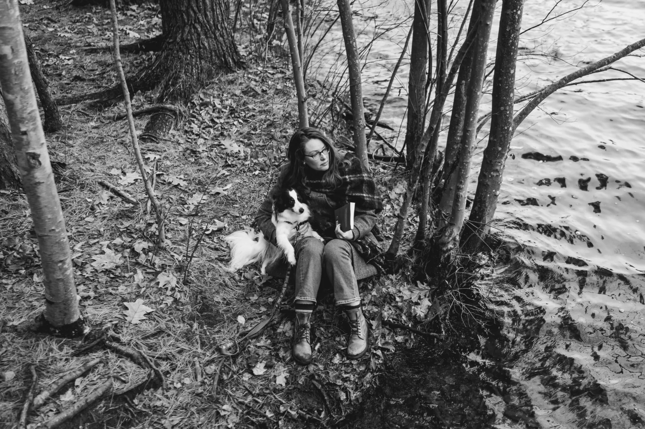 Woman sitting under birch trees with a small dog beside a lake