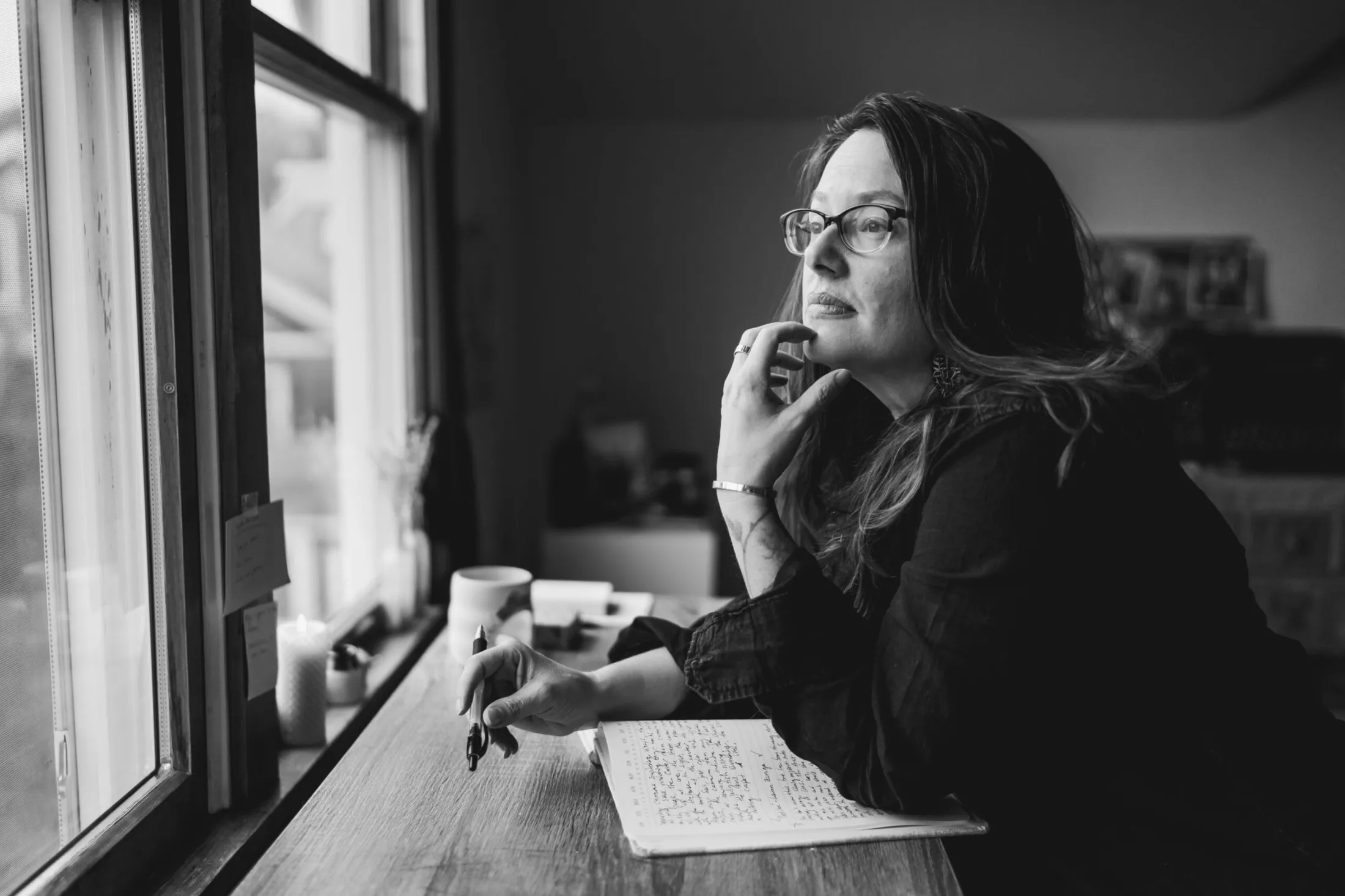 Woman in glasses gazing out a window at a desk with a notebook