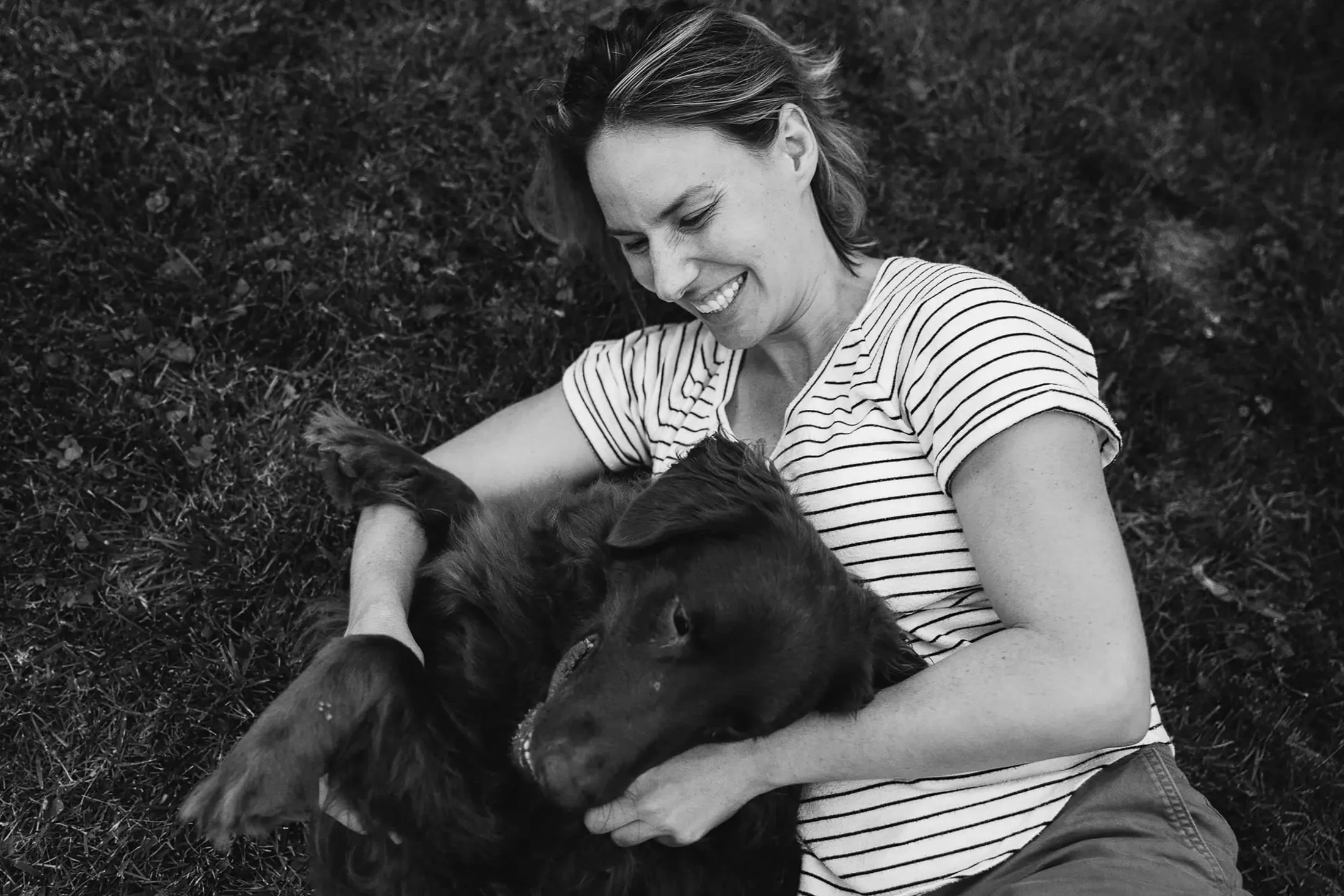 Woman laughing as she hugs a black dog on the grass