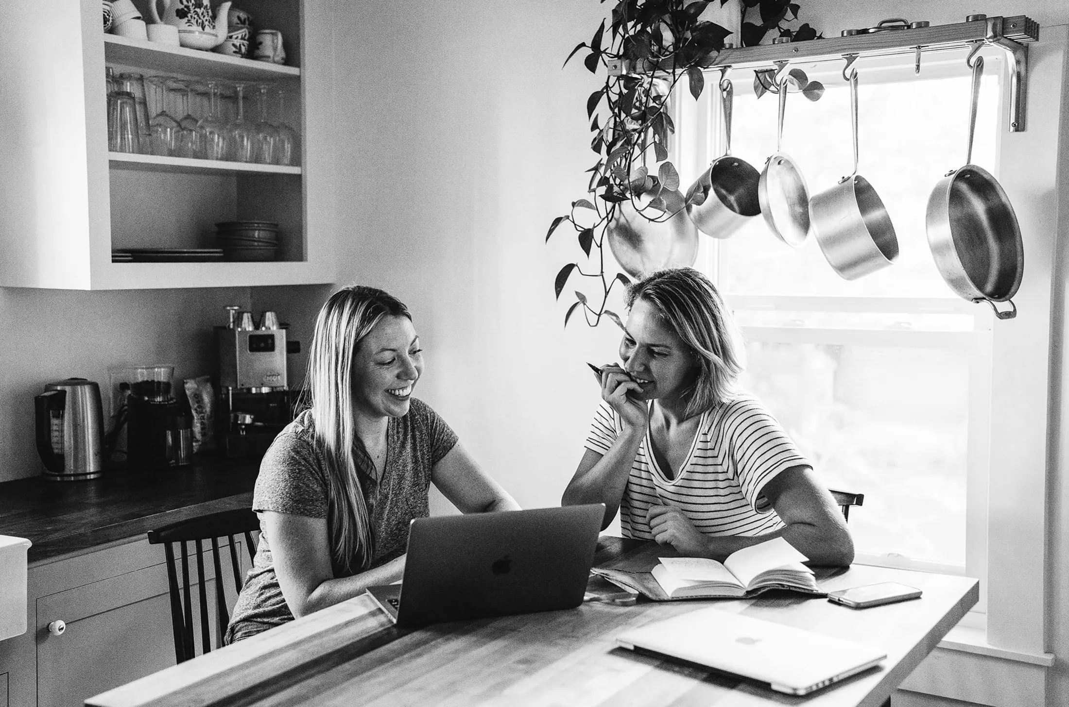 Two women laughing at a kitchen table with a laptop under hanging pots