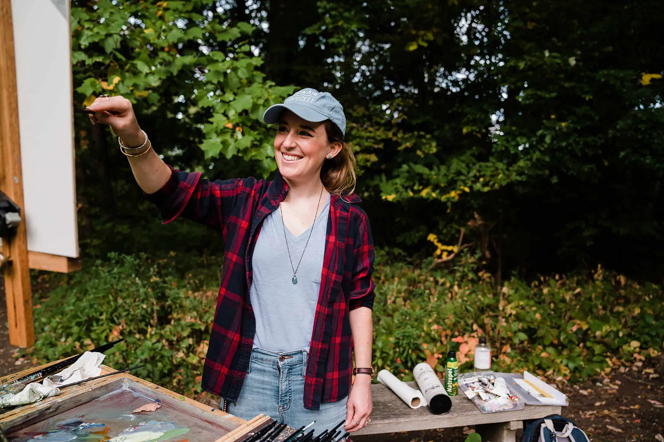 Woman in a plaid shirt and cap gesturing beside an outdoor painting table