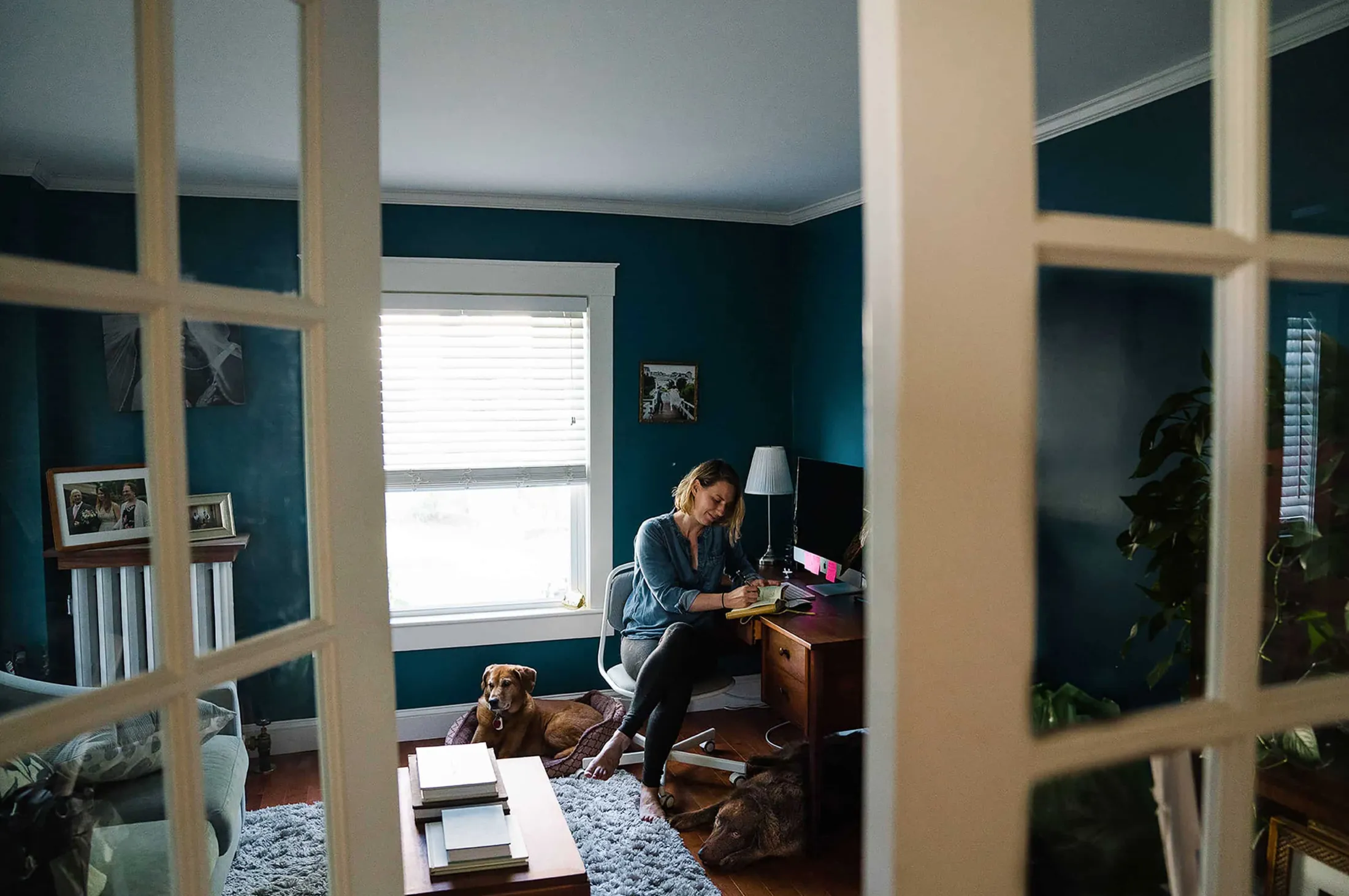 Woman working at a desk in a teal study with a dog, seen through French doors