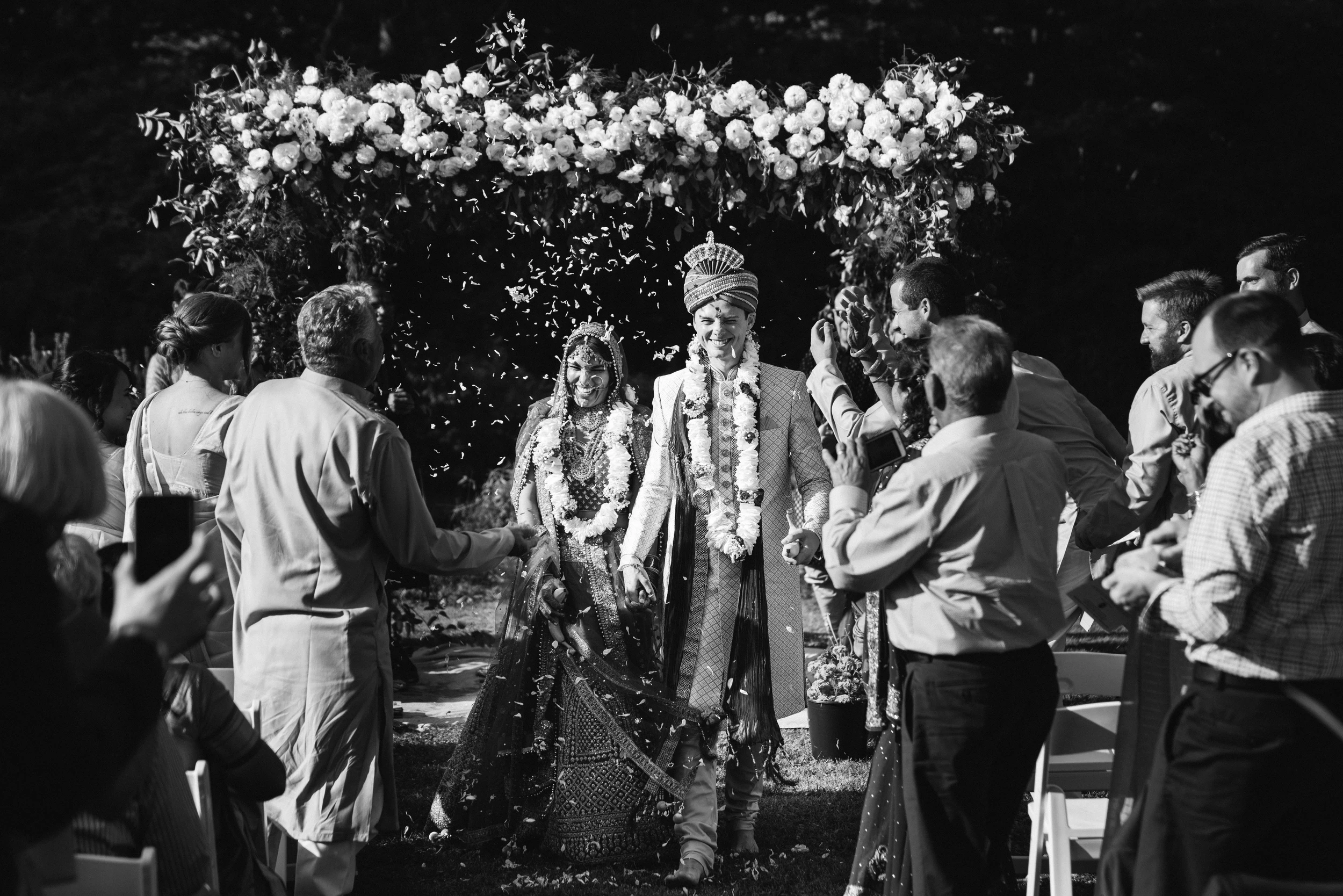 A couple under a flower-covered arch as guests crowd in around them
