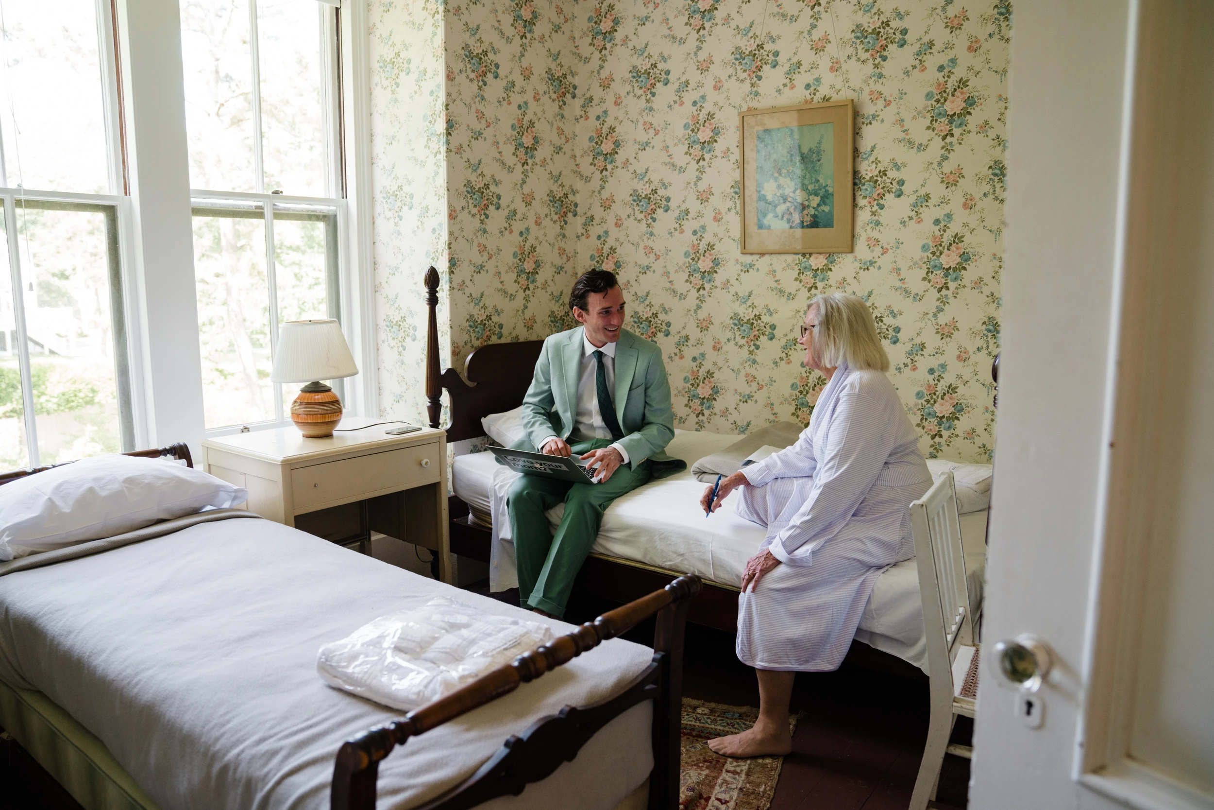 A man in a green suit talks with a woman in a robe in a room with floral wallpaper and two single beds