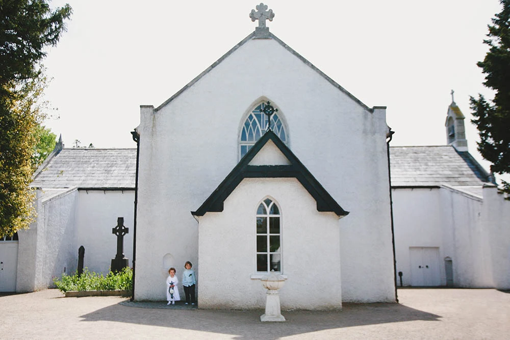 A small white church with a cross on its gable, the couple tiny at the door