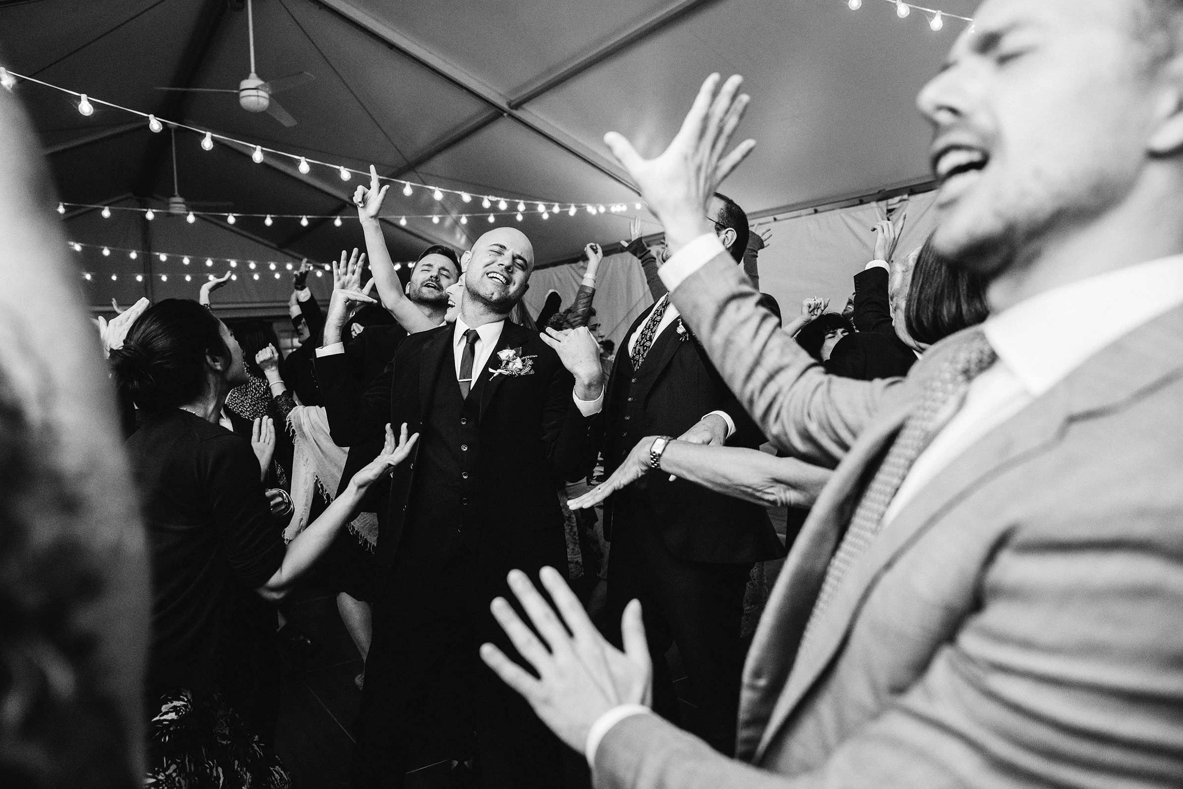 Men singing with their arms in the air under string lights