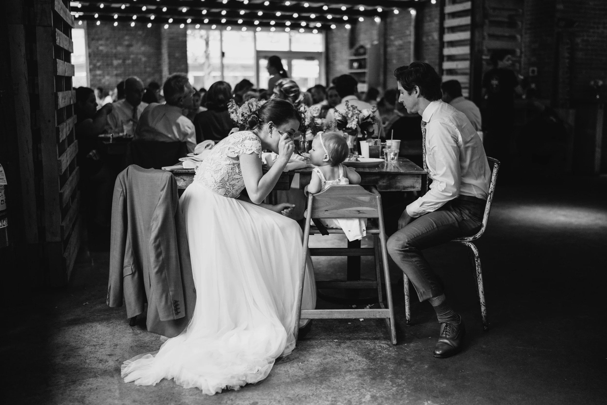 Bride leans in to feed a toddler in a high chair at a long reception table under string lights
