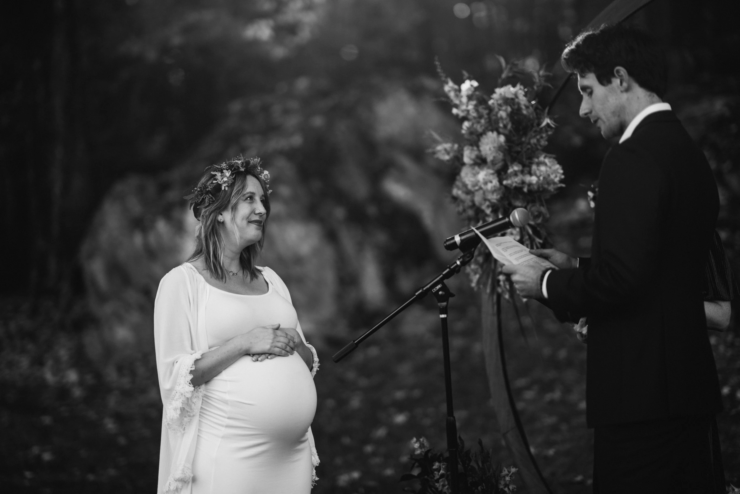 Pregnant bride in a flower crown smiles at the groom reading vows into a microphone