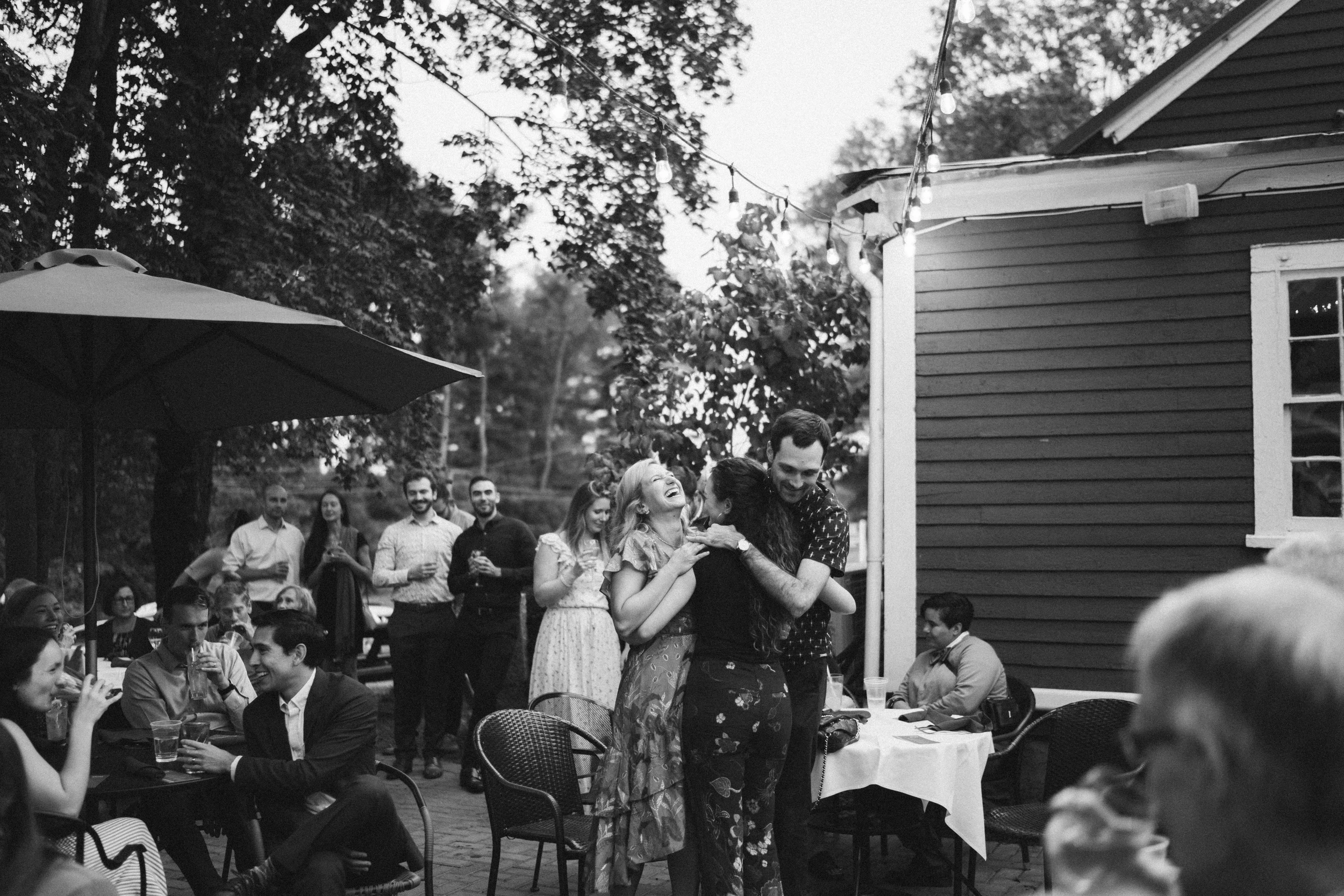 Guests gathered on a patio beside a grey house under the trees