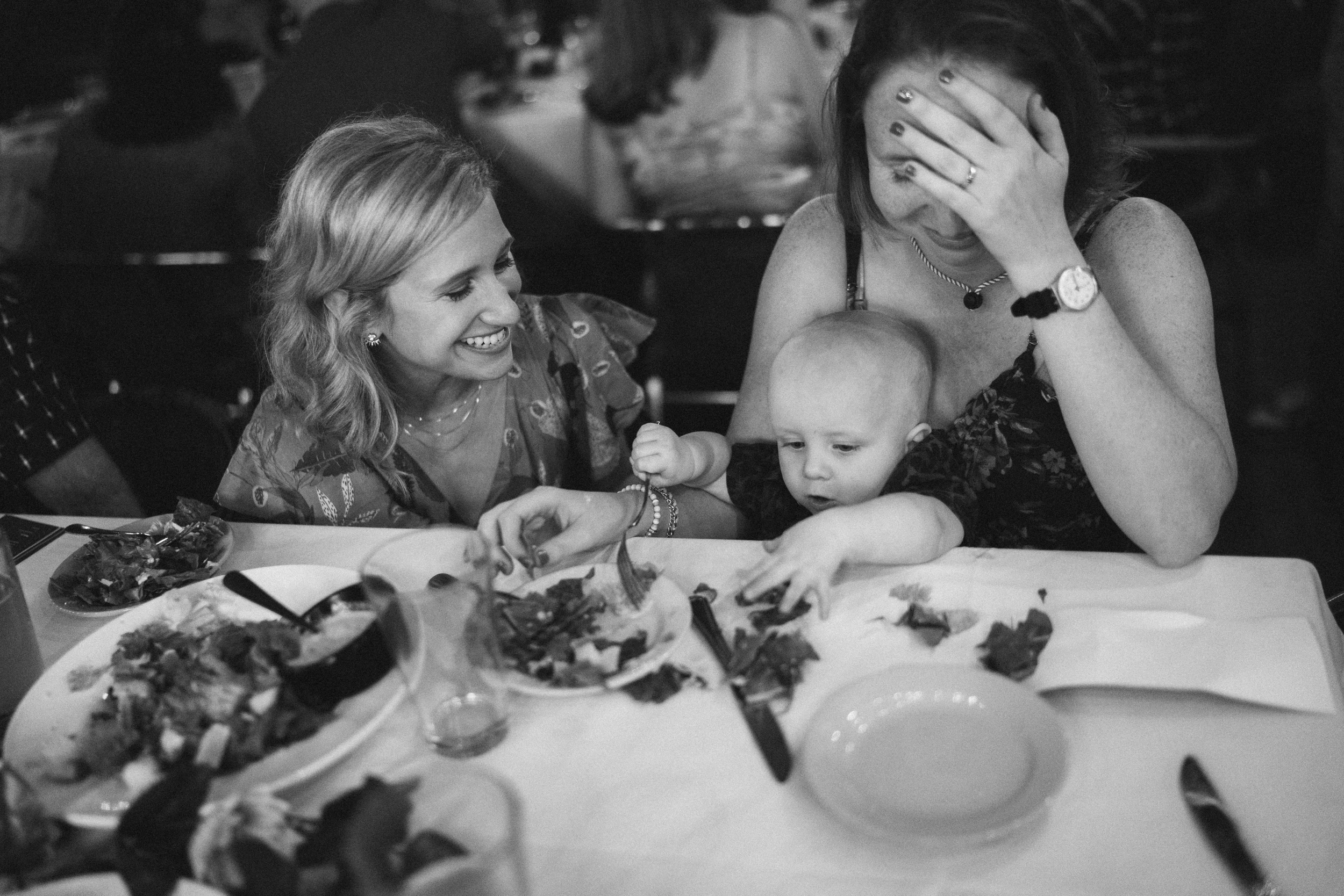 A woman laughing at the table with a baby reaching for a plate