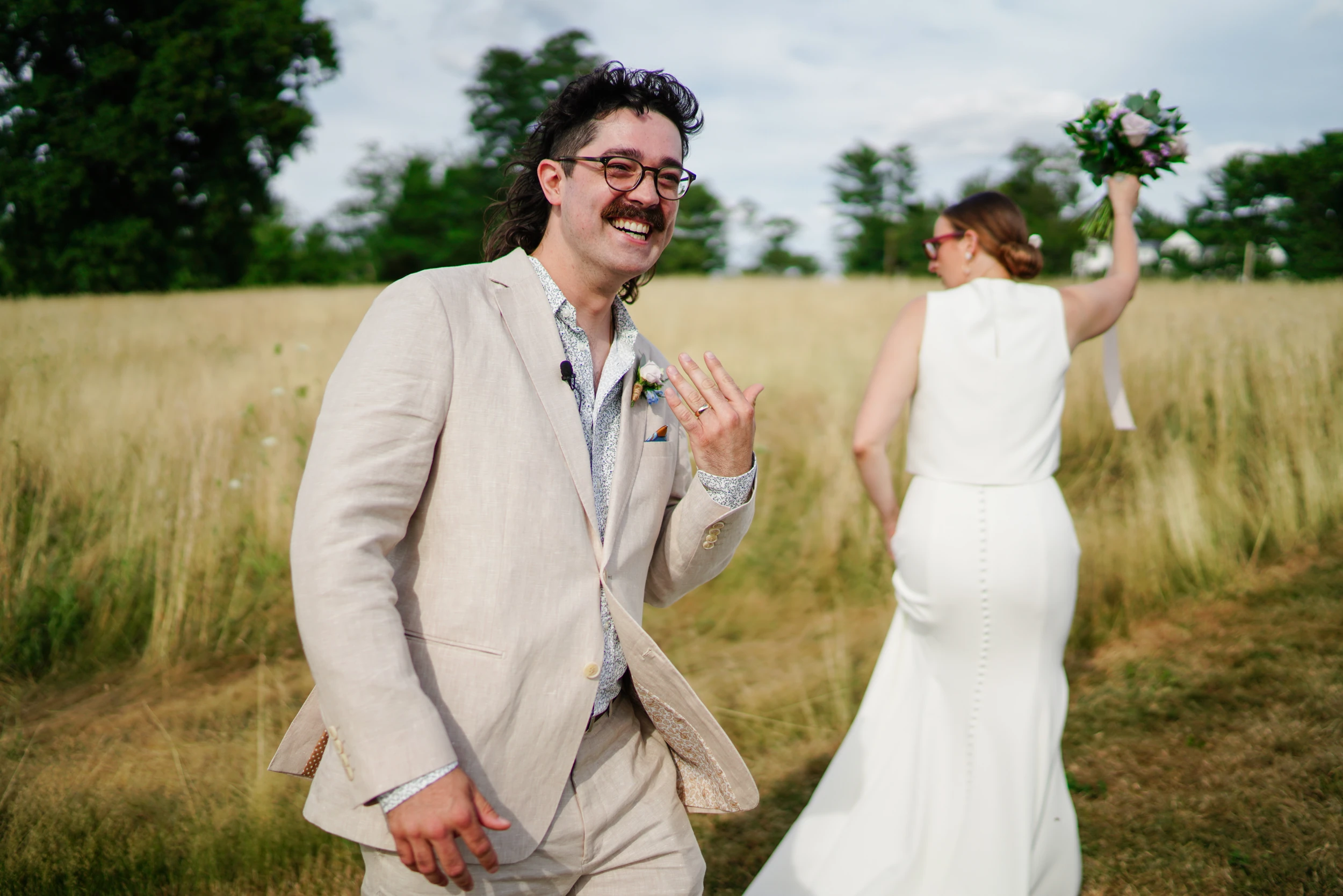 Groom grinning and showing his ring hand while the bride walks off raising her bouquet in a tall-grass field