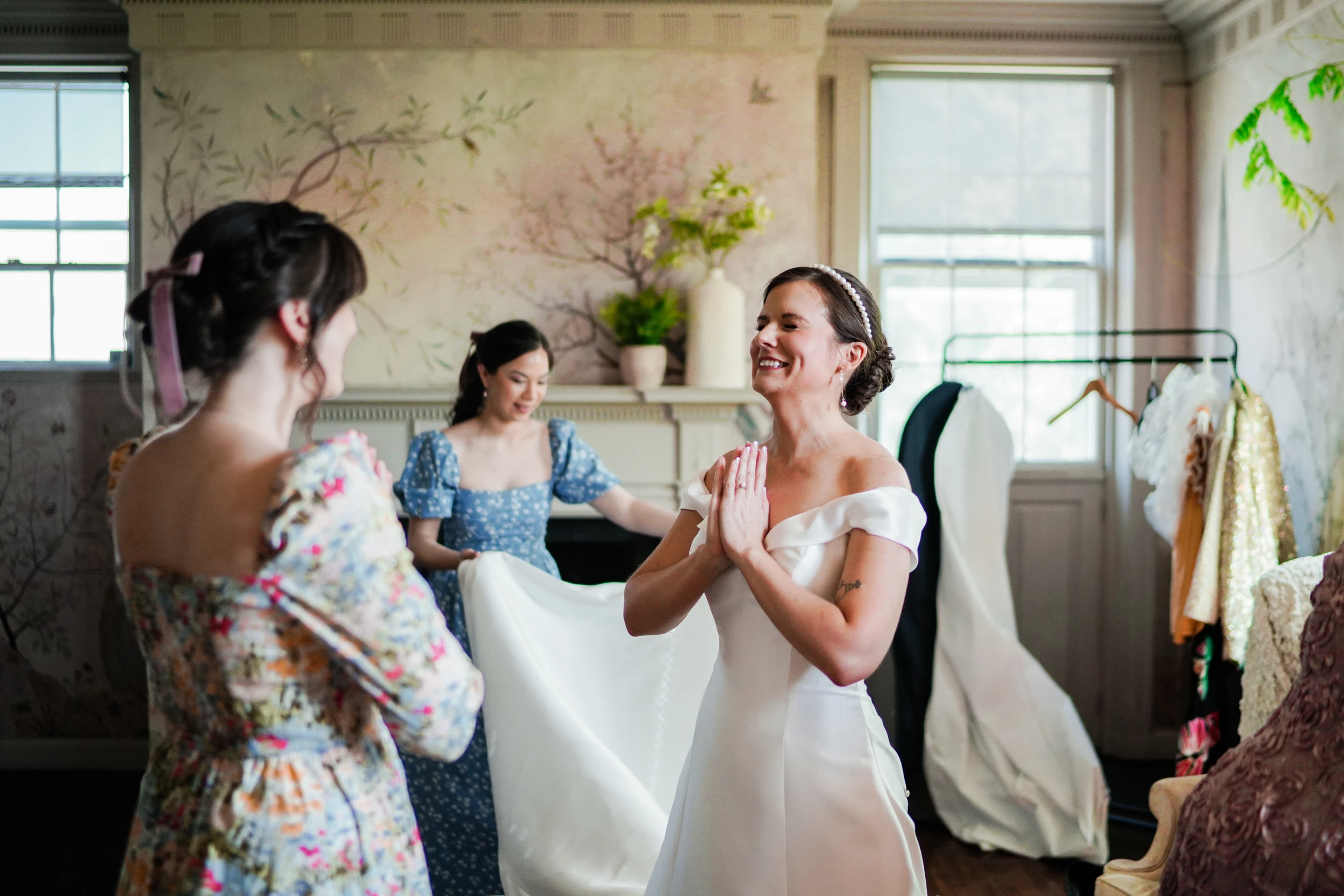 Bride with hands pressed together laughing as two women lift her train in a room with painted wallpaper