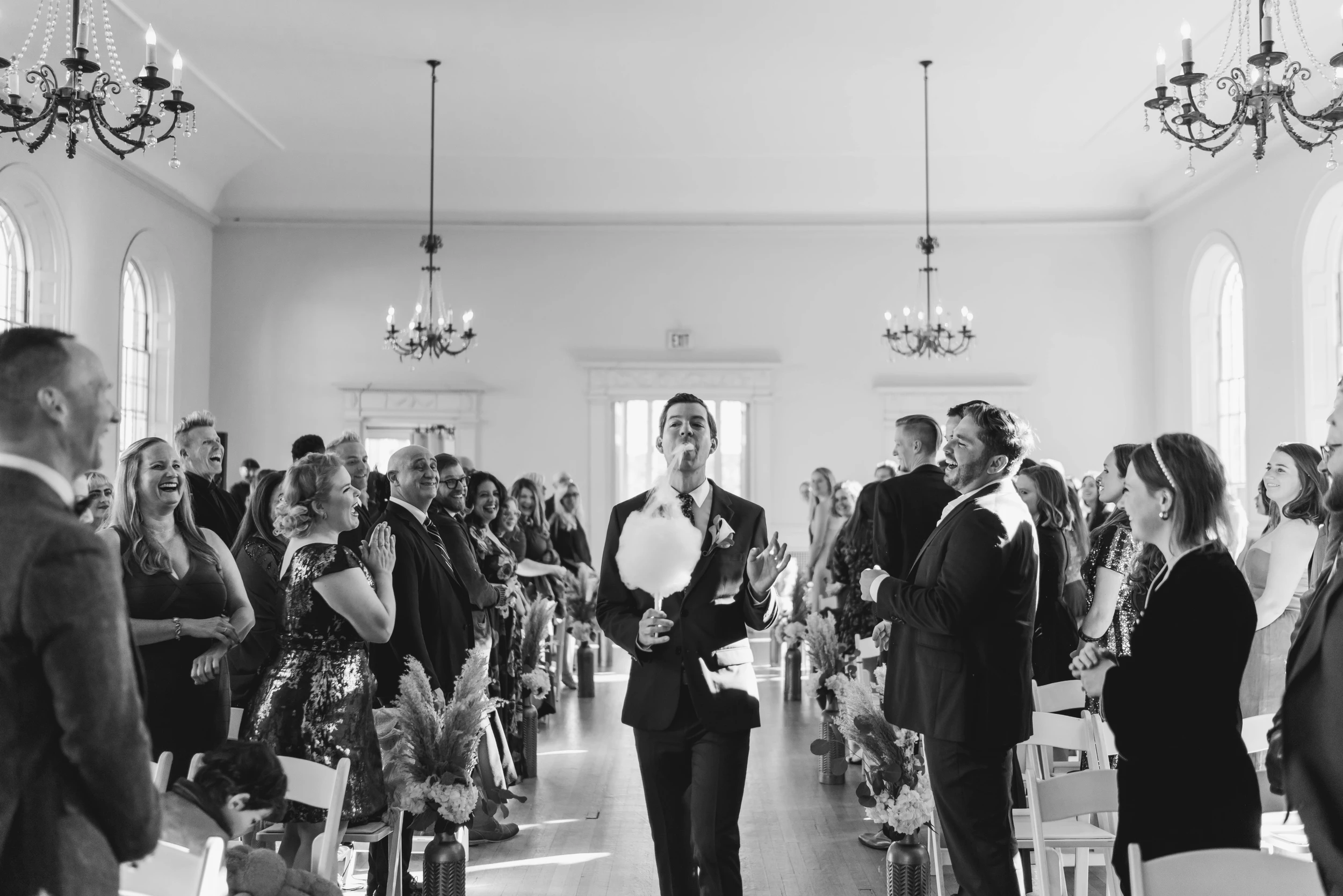 Guests standing along the aisle of a white hall under chandeliers