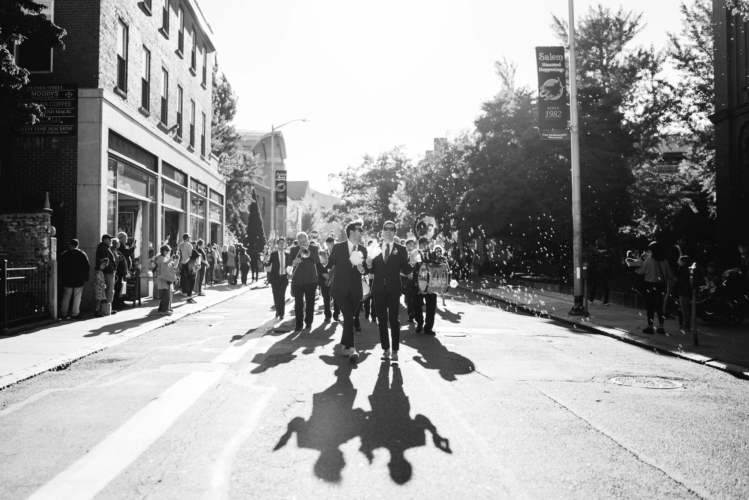 Two grooms lead a brass band procession down a sunlit street, long shadows and bubbles