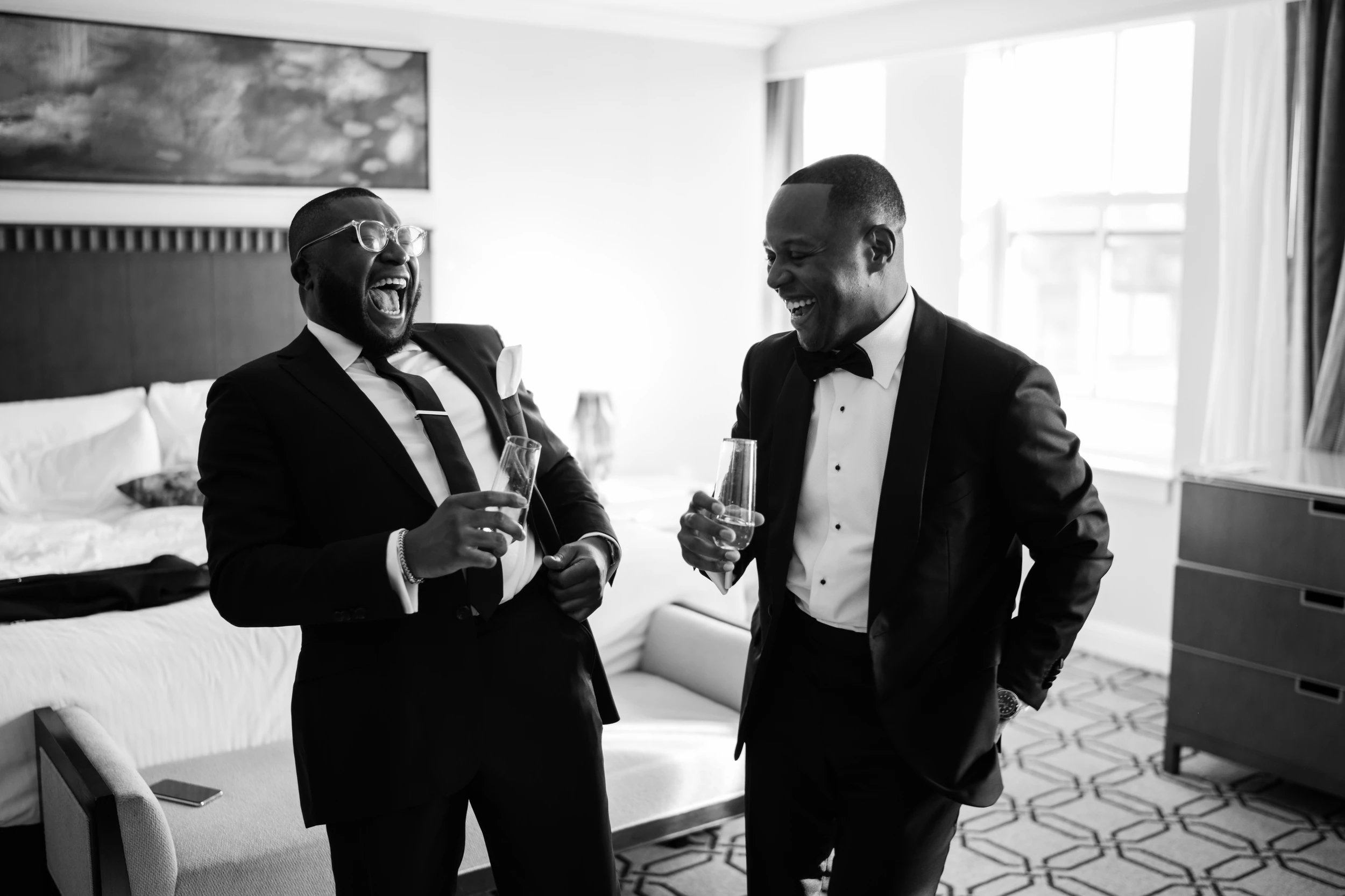 Two men in tuxedos laughing with champagne flutes in a hotel room